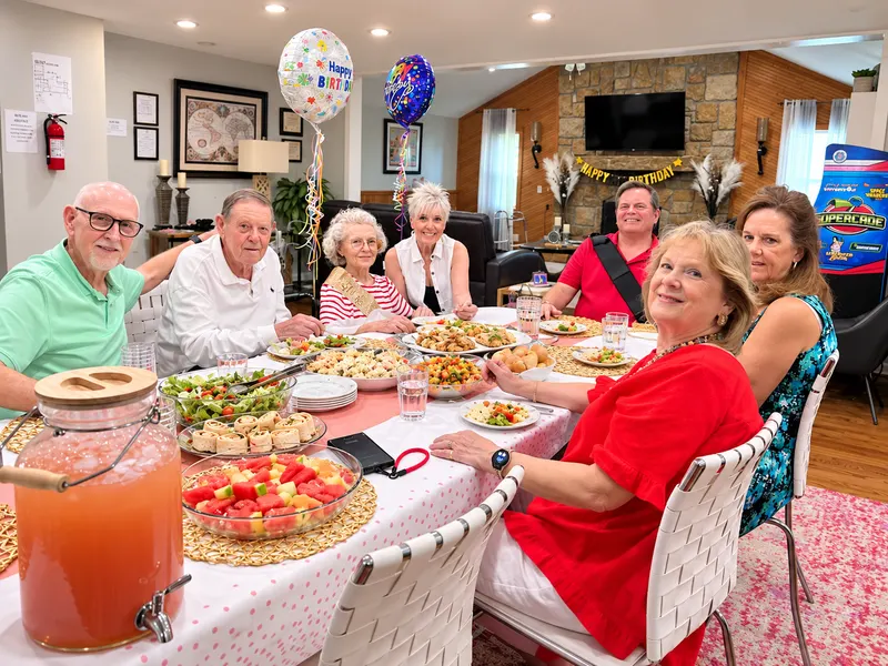 Residents and family around a long table set for a birthday lunch, with balloons in the background.